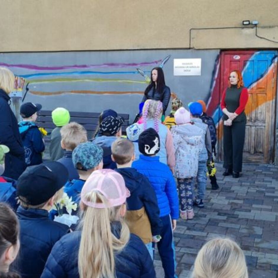 Atvērto durvju dienas dalībnieki ārā pie mākslas skolas, klausās mākslas skolotājā