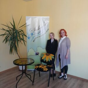 Skolotāja Ilze Šuca ar skolnieci un viņas diplomdarba aizstāvēšanas darbiem (krēsls un galds ar saulespuķēm)