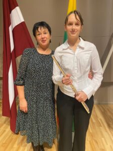 Jauna sieviete smaidot tur galda spēli ar vāku, uz kura redzams teksts "Flow", melns kaķis un zelta retrīvers, kā arī diplomu ar tekstu: "DIPLOMS Alissai Liepai par iegūto I vietu Pašaminstumentu spēles konkurss BALTU PUTNI 2023".