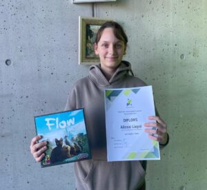 Jauna sieviete, smaidot kamerā, tur galda spēles kasti ar nosaukumu "Flow", uz kuras attēlots melns kaķis, suns un pilsētas ainava, un diplomu ar tekstu: "Polaminstrumentu spēles konkurss "BALTU PUTNI 2020" DIPLOMS Alissai Liepai (Rīgas Centra humanitārā vidusskola) par iegūto I vietu. 2020. gada 2. marts.