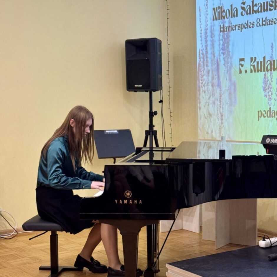 Jauna sieviete tumši zaļā blūzē un melnos svārkos spēlē melnu Yamaha flīģeli uz koka parketa grīdas. Fonā uz ekrāna redzams uzraksts 