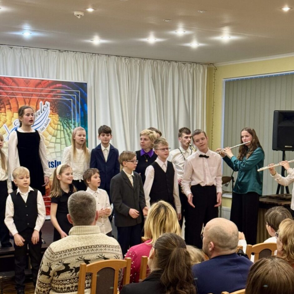 Bērnu un jauniešu grupa uzstājas koncertā, daži dzied, citi spēlē flautas. Priekšplānā sēž publika. Aizmugurē redzams krāsains plakāts ar tekstu, kas daļēji salasāms kā 