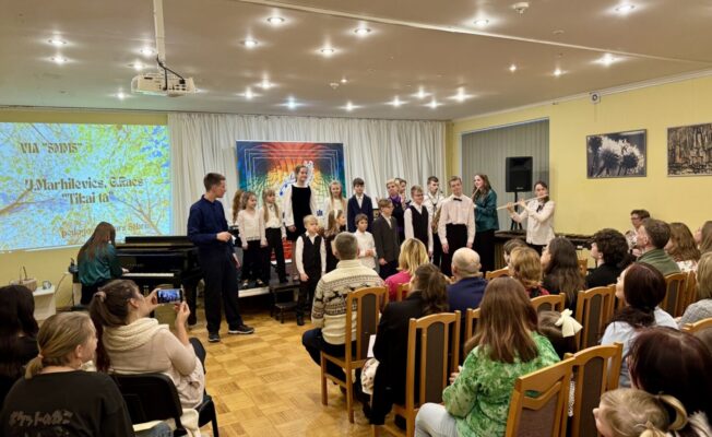 Bērnu mūzikas priekšnesums: uz skatuves stāv bērni un jaunieši kopā ar diriģentu, kamēr priekšplānā sēž pieaugušo auditorija. Uz projektora ekrāna redzams teksts: 