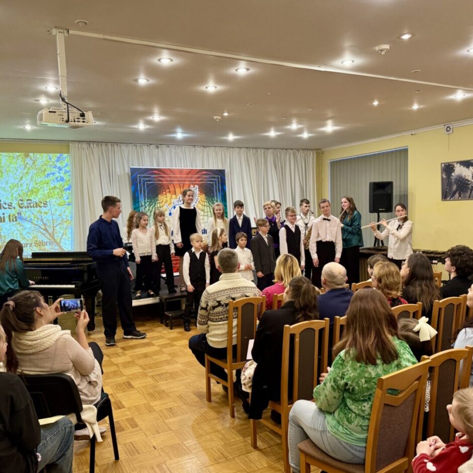 Bērnu mūzikas priekšnesums: uz skatuves stāv bērni un jaunieši kopā ar diriģentu, kamēr priekšplānā sēž pieaugušo auditorija. Uz projektora ekrāna redzams teksts: 
