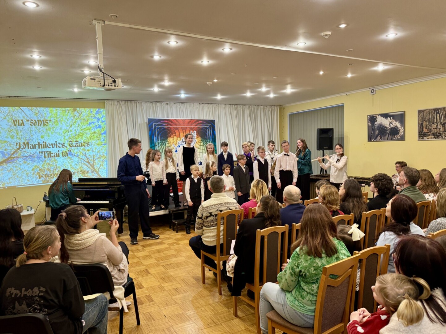 Bērnu mūzikas priekšnesums: uz skatuves stāv bērni un jaunieši kopā ar diriģentu, kamēr priekšplānā sēž pieaugušo auditorija. Uz projektora ekrāna redzams teksts: 