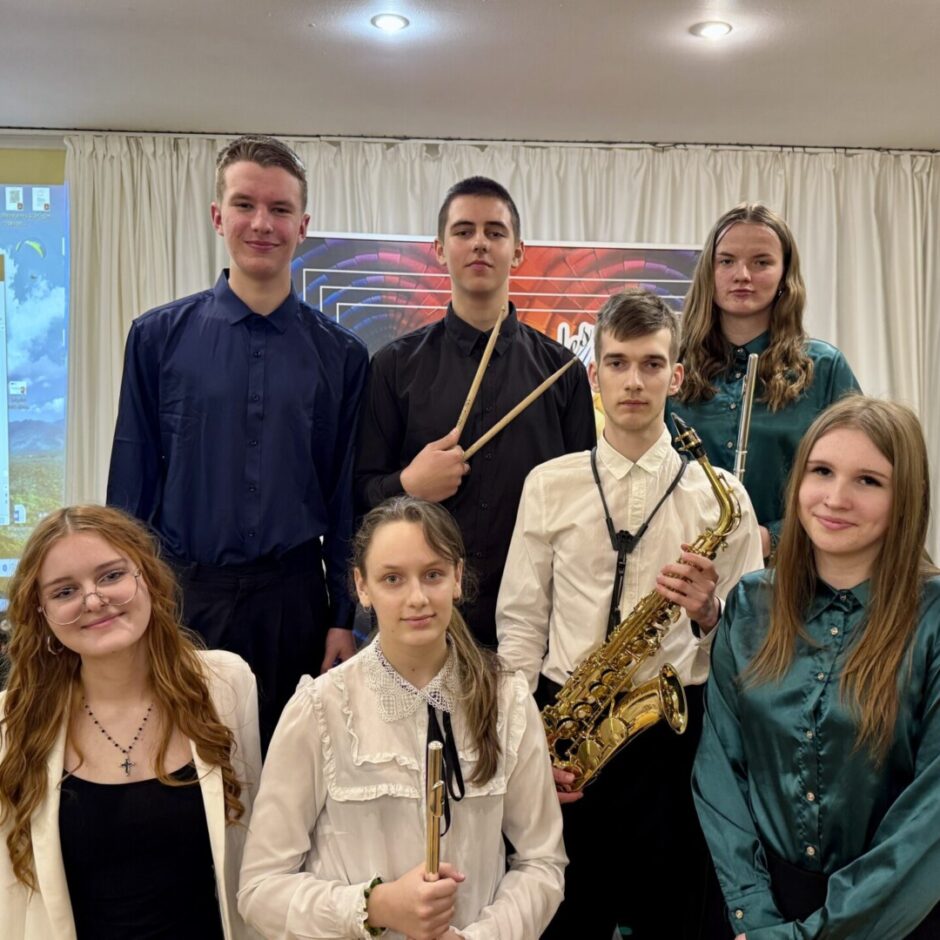 Septiņi jaunieši (četri puiši un trīs meitenes) pozē grupas fotogrāfijai. Viens puisis tur bungas vālītes, cits tur saksofonu, viena meitene tur flautu un otra tur nelielu pūšaminstrumentu. Fonā ir balts aizkars, projektora ekrāns ar tekstu 