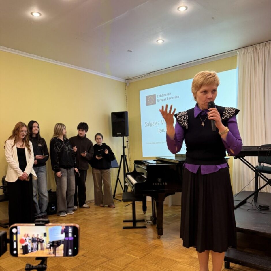 Sieviete ar mikrofonu runā, blakus melnas klavieres un jauniešu grupa. Aiz muguras uz projekcijas ekrāna redzams uzraksts 