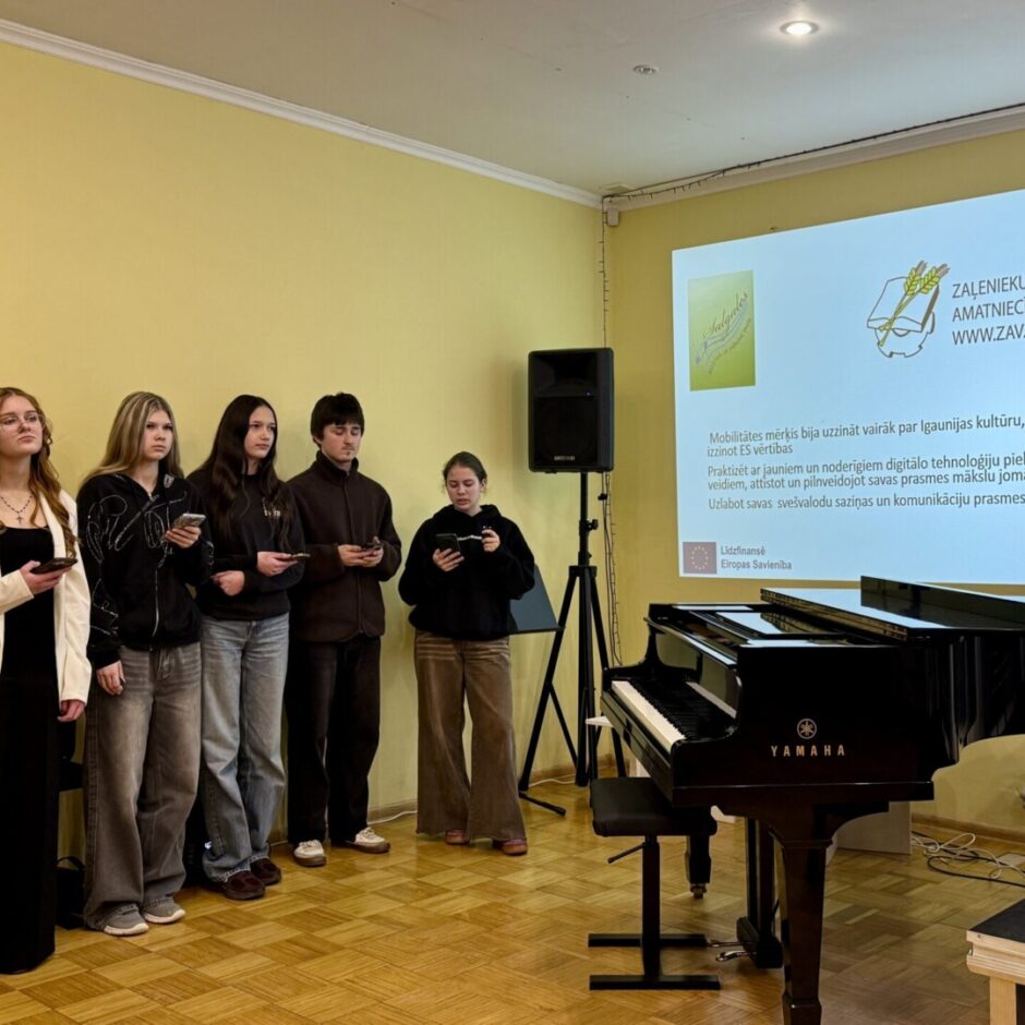 Pieci jaunieši stāv rindā, turot mobilos tālruņus. Uz dzeltenas sienas tiek projicēta prezentācija ar virsrakstu 