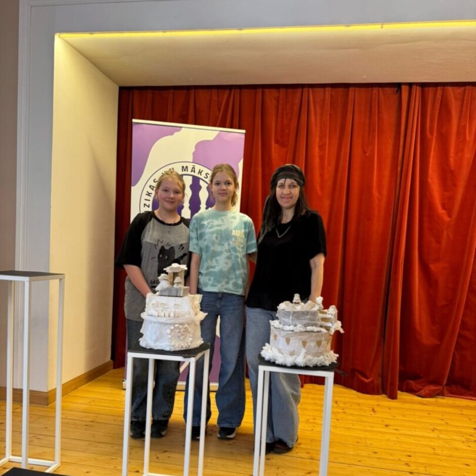 Divas meitenes un sieviete stāv uz koka skatuves, smaida. Viņu priekšā uz baltiem statīviem atrodas divas daudzlīmeņu dekoratīvas kūkas. Aizmugurē ir sarkani aizkari un violets reklāmkarogs ar tekstu 