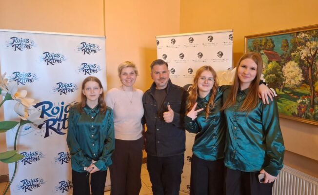 Pieci cilvēki – trīs jaunas meitenes vienādos tirkīzzilos kreklos, sieviete un vīrietis – pozē fotogrāfijai. Vīrietis rāda īkšķi uz augšu, bet viena meitene rāda miera zīmi. Fonā ir stendi ar uzrakstiem 