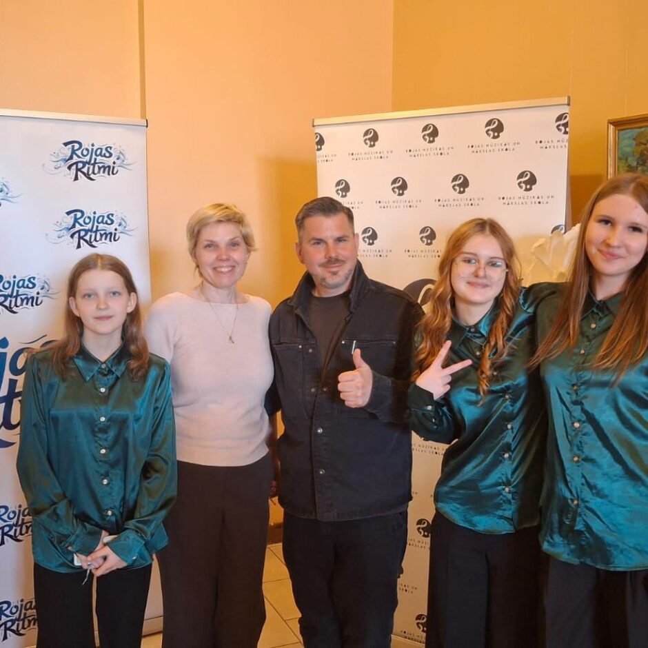 Pieci cilvēki – trīs jaunas meitenes vienādos tirkīzzilos kreklos, sieviete un vīrietis – pozē fotogrāfijai. Vīrietis rāda īkšķi uz augšu, bet viena meitene rāda miera zīmi. Fonā ir stendi ar uzrakstiem 