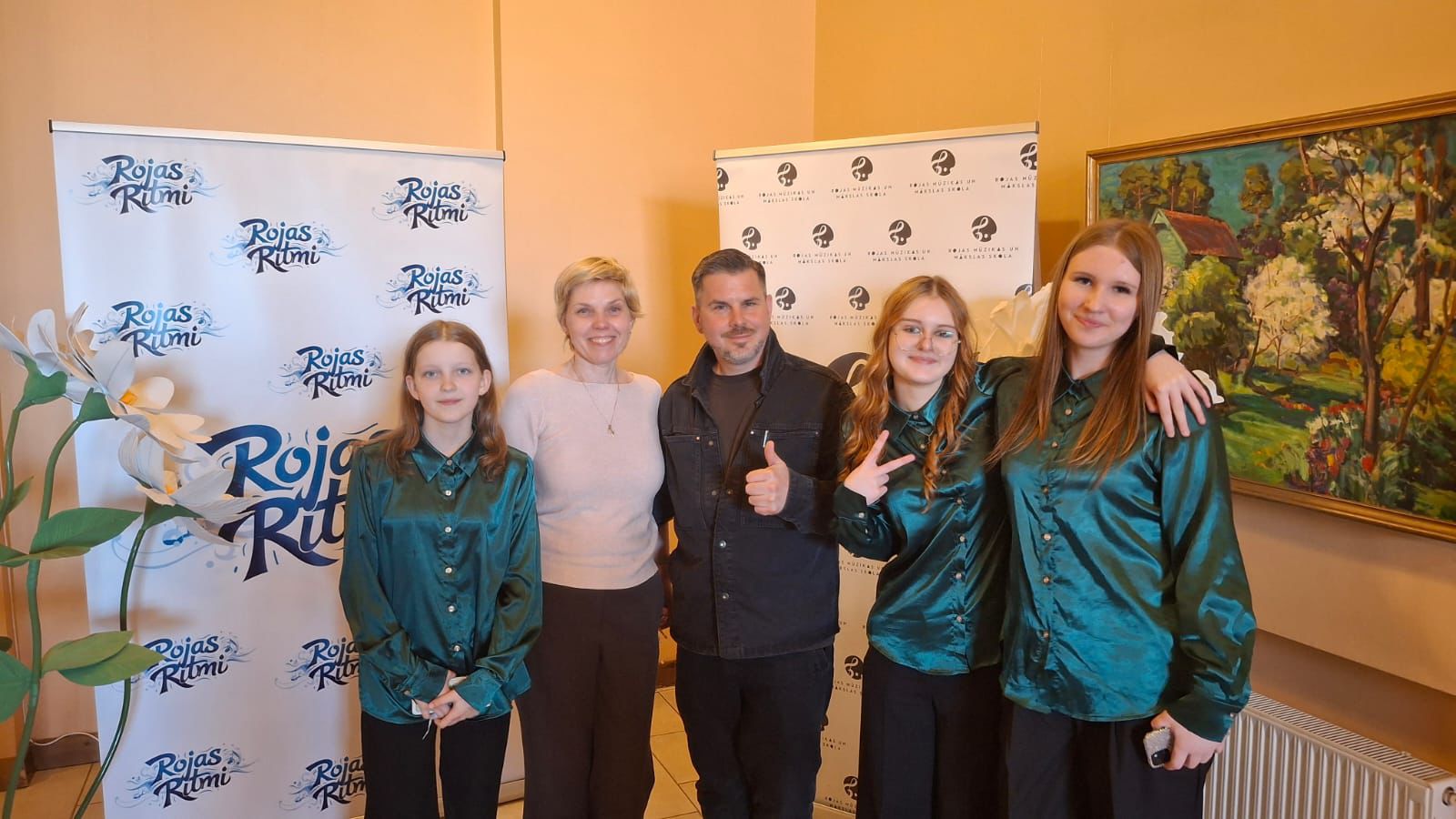 Pieci cilvēki – trīs jaunas meitenes vienādos tirkīzzilos kreklos, sieviete un vīrietis – pozē fotogrāfijai. Vīrietis rāda īkšķi uz augšu, bet viena meitene rāda miera zīmi. Fonā ir stendi ar uzrakstiem 