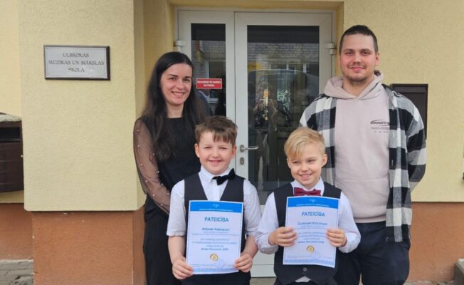 Sieviete, vīrietis un divi zēni smaida, stāvot pie ēkas ar uzrakstu 