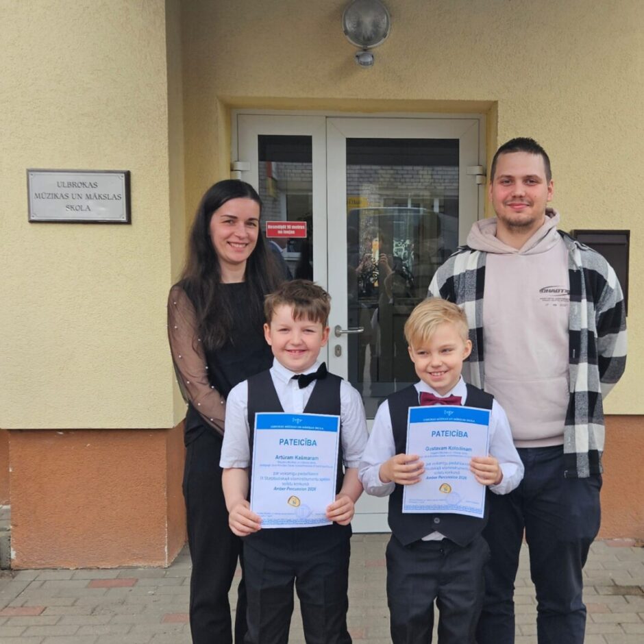 Sieviete, vīrietis un divi zēni smaida, stāvot pie ēkas ar uzrakstu 