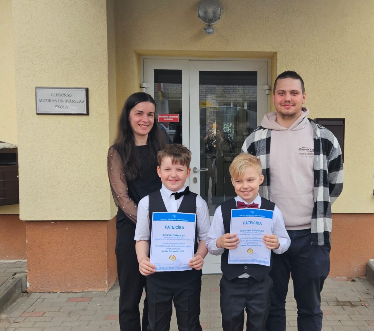 Sieviete, vīrietis un divi zēni smaida, stāvot pie ēkas ar uzrakstu 